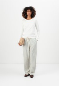 Haut blanc à manches longues avec un encolure ronde, associé à un pantalon large à rayures claires. Le mannequin tient un sac beige et porte des chaussures marron.