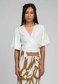 Femme aux cheveux tressés portant un haut court enroulé blanc et une jupe à motifs marron et blancs, se tenant devant un fond gris uni.