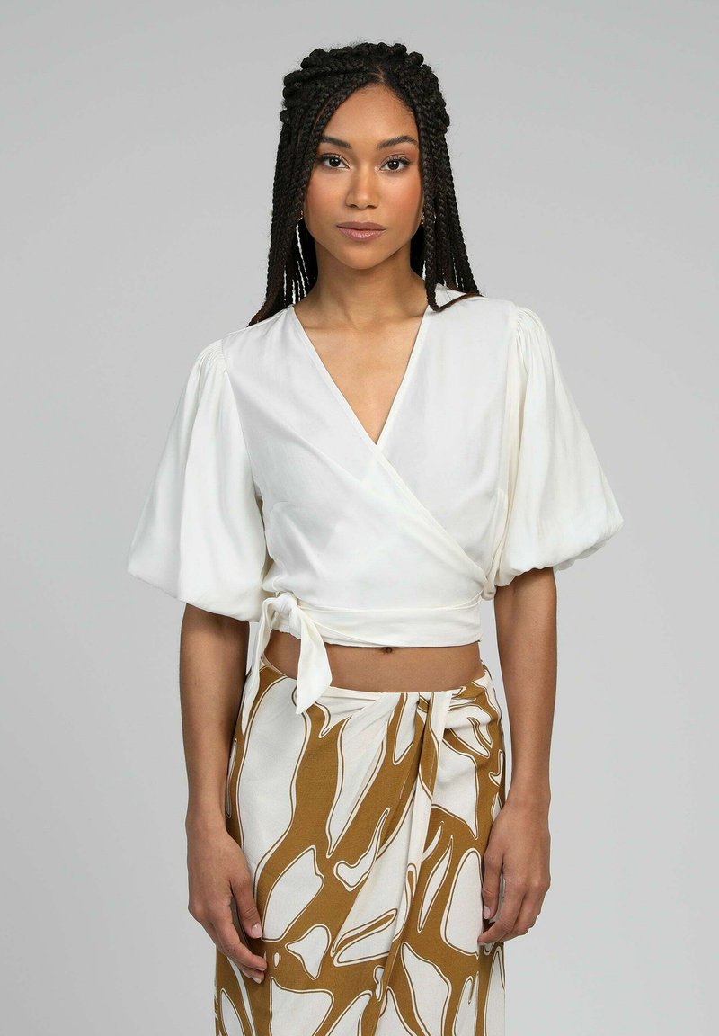 Femme aux cheveux tressés portant un haut court enroulé blanc et une jupe à motifs marron et blancs, se tenant devant un fond gris uni.