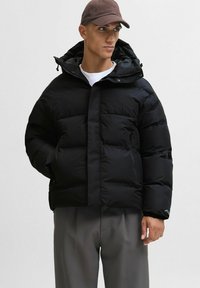 Giacca a vento nera con una finitura leggermente lucida, zip anteriore, colletto alto e cappuccio regolabile. Presenta tasche con zip e una vestibilità rilassata.