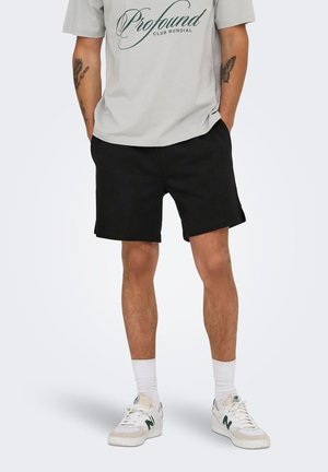 Mand iført lysegrå "Profound Club Mundial" t-shirt, sorte shorts, hvide sokker, hvide New Balance-sneakers, med tatoveringer på underarme og hænderne i lommerne.