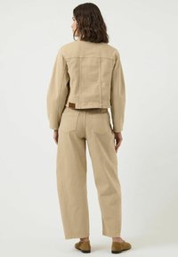 Vrouw met schouderlang bruin haar draagt een bijpassende beige spijkerjack en wijde spijkerbroek, staande met de rug naar de camera tegen een effen achtergrond.