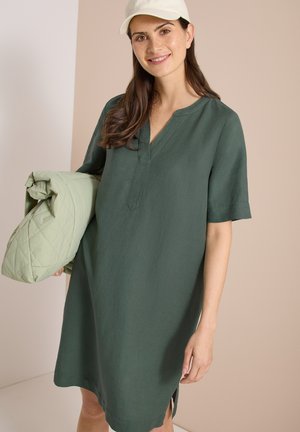 Kvinde iført en mørkegrøn kortærmet kjole og hvid kasket, der holder en lysegrøn quiltet jakke og smiler mod en beige og hvid baggrund.