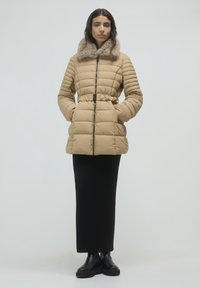 Donna che indossa una giacca trapuntata beige con colletto in pelliccia, gonna lunga nera e stivali neri, in piedi con le mani nelle tasche della giacca.