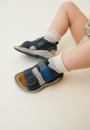 Sandalete din plasă albastre cu curea velcro, talpă din cauciuc gri și vârf portocaliu. Asortate cu șosete albe cu dungi gri pe picioare mici.