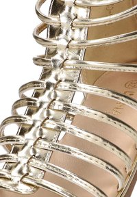 Sandalia de tiras en oro metálico que presenta una serie de tiras curvas entrelazadas. Textura suave con suela plana y un interior de color beige claro.