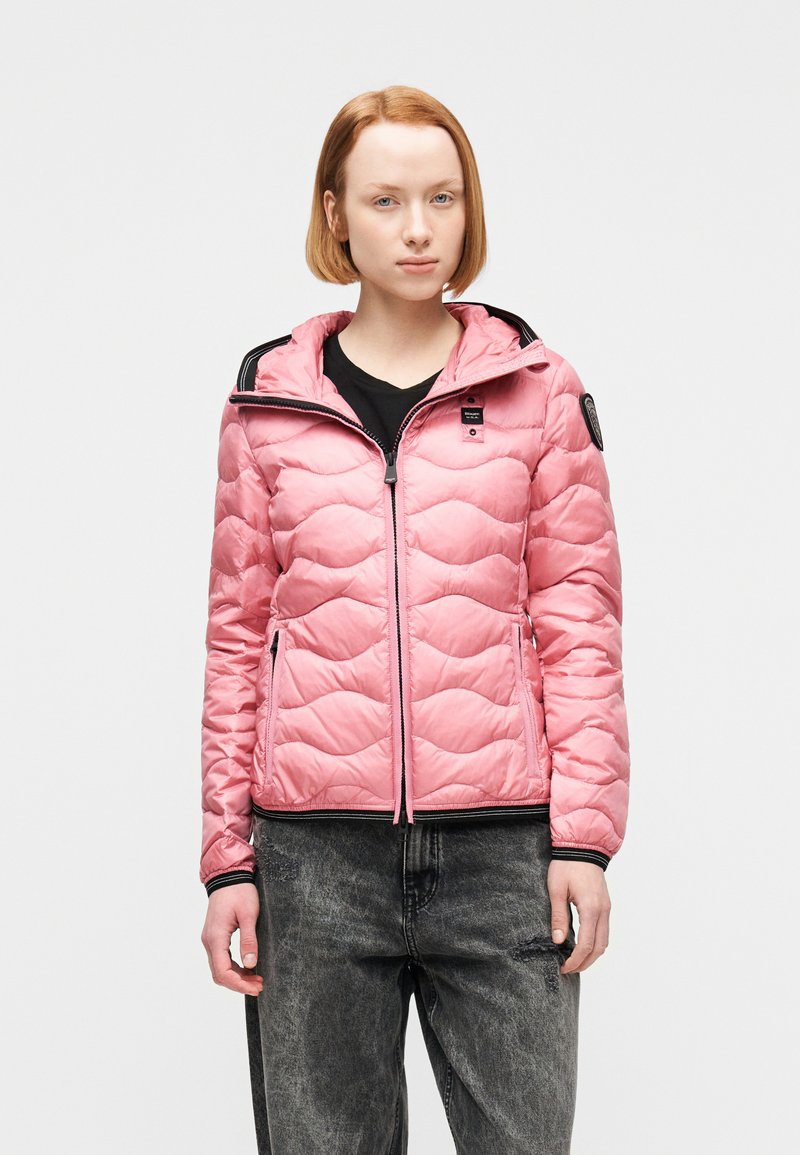 Νεαρή γυναίκα με κοντά κόκκινα μαλλιά φοράει ροζ μπουφάν με επένδυση πάνω από μαύρο πουκάμισο και φαρδιά σκούρα γκρι τζιν, στέκεται μπροστά σε λευκό φόντο.