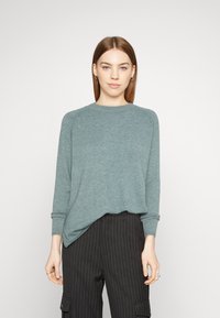Pullover in maglia verde menta con scollo a girocollo, maniche lunghe e vestibilità rilassata. Indossato su pantaloni neri a righe. Texture liscia, design minimal.