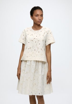 Kobieta ubrana w biały top z krótkim rękawem z ażurowej koronki oraz pasującą spódnicę do kolan, stojąca na jednolitym jasnym tle.