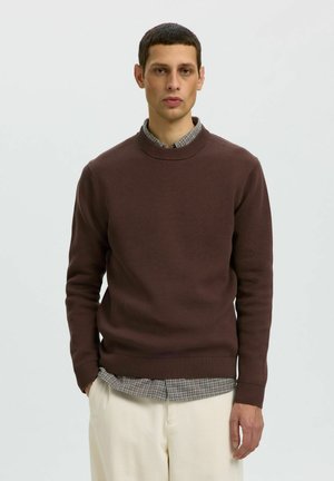 Homme portant un pull en maille marron par-dessus une chemise à carreaux grise et un pantalon en velours côtelé crème, debout devant un fond clair uni.