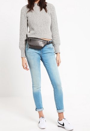 Femme portant un jean bleu clair retroussé, un pull en maille gris, des baskets blanches Nike et une banane noire métallique à la taille sur fond blanc.