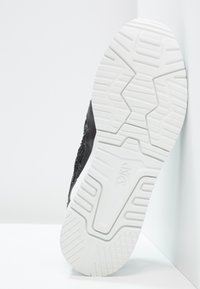 Svart sneaker med en strukturerad ovandel och vit gummisula. Sulan har ett mönstrat mönster för bra grepp och stabilitet.