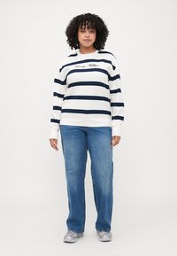 Stripet langermet sweatshirt i hvit og marine, med rund hals og brodert logo. Et par blå jeans og grå sneakers.