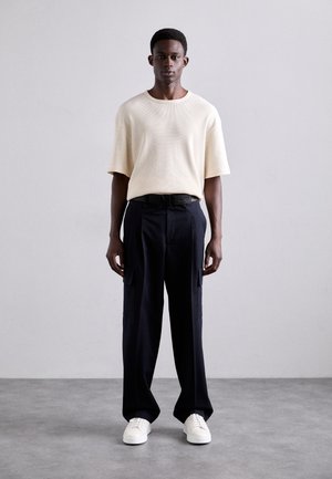 Šviesiai smėlinė megzta trumpa rankovė, laisvas kirpimas, derinama su juodomis cargo kelnėmis su šoninėmis kišenėmis ir baltais sportbačiais. Neutralus fonas.