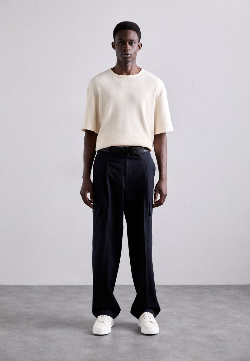 Gai bēšā tamborētā īsām piedurknēm krekls, brīvs griezums, apvienots ar melniem cargo biksēm ar sānu kabatām un baltām sporta kedām. Neitrāls fons.