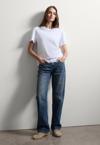 T-shirt bianco a maniche corte abbinato a jeans in denim blu dalla vestibilità ampia. Il modello è in piedi contro un muro chiaro, con le mani in tasca, indossando scarpe beige.