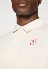 Beige polo shirt med krave, der har en knaplukning og et rødt og hvidt broderet løve-logo på venstre bryst. Glat stofstruktur.