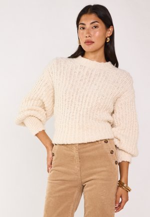 Maglione lavorato a maglia color crema con una trama a coste e maniche ampie, abbinato a pantaloni in velluto a coste color cammello con dettagli sui bottoni.