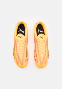 Zapatos deportivos amarillos brillantes con acentos rosas, un diseño de parte superior texturizado y plantillas negras con un logo blanco. Los cordones están exhibidos de manera prominente.