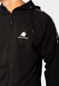 Zwarte ritshoodie van een gestructureerde stof, met een wit logo op de borst en branding langs de mouwen. Verstelbare trekkoordcapuchon.