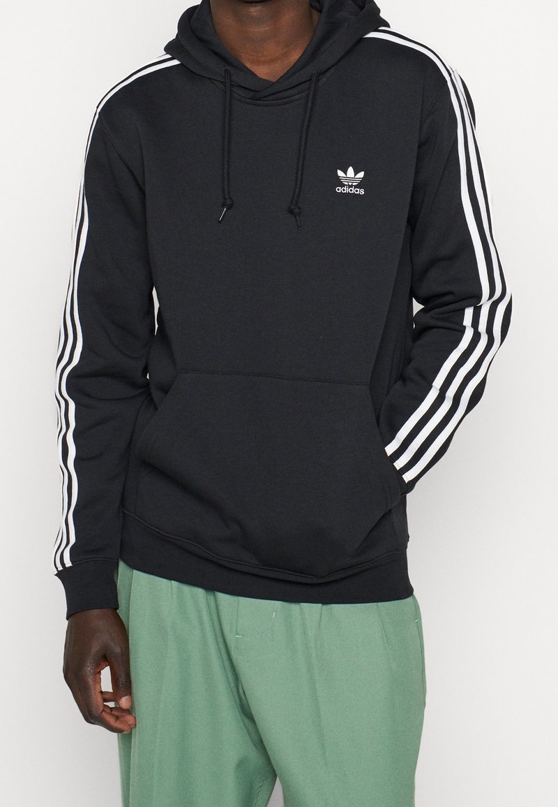 Schwarzer Hoodie mit Kapuze und Kordelzug, weißen drei Streifen-Design an den Ärmeln und kleinem weißen Adidas-Logo auf der Brust. Material aus Baumwollmischung.