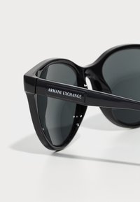 Juodos acetato saulės akiniai su tamsiais objektyvais, turintys geometrinį rėmelio dizainą ir "Armani Exchange" logotipą ant šventyklos.
