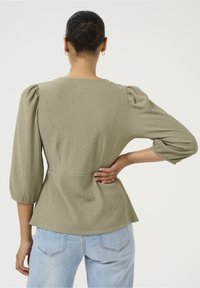 Kvinde med hår i en knold, iført en tekstureret olivengrøn bluse med puffærmer og lyseblå jeans, stående med hånden på lænden.
