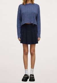 Zkrácený modrý svetr s pleteným vzorem a dlouhými rukávy v kombinaci s černou skladanou sukní. Modelka má na sobě bílé kotníkové ponožky a robustní černé polobotky.