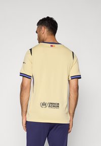 Bež sportski dres s crnim i ljubičastim detaljima, s logotipom na leđima i malim detaljem zastave na vratu.