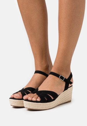 Sandali wedge in suede nero con una piattaforma in juta intrecciata, caratterizzati da un design a strisce incrociate e una cinghia alla caviglia con dettagli in metallo.