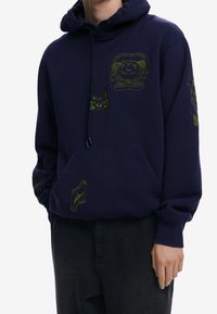 Granatowy polar z bawełny, ozdobiony jasnobrązowymi grafikami, z kieszenią kangurką i kapturem z troczkami. Tekst i wzory na rękawach.