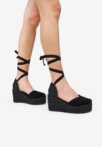 Black wedge espadrilles featuring a soft suede upper, textured side panels, and crisscross ankle straps, with a braided jute midsole.