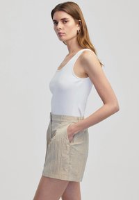 Witte geribbelde tanktop gecombineerd met lichtbeige textuuroveralls met zijzakken en een ceintuur in de taille. Staande pose tegen een effen achtergrond.