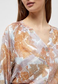 Blusa con bottoni caratterizzata da un motivo astratto in arancione, blu e marrone su uno sfondo chiaro. Tessuto morbido con vestibilità rilassata.