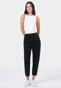 Zwarte corduroy tapered broek met voorzakken, tentoongesteld met een aansluitend wit mouwloos topje en witte sneakers, tegen een neutrale achtergrond.