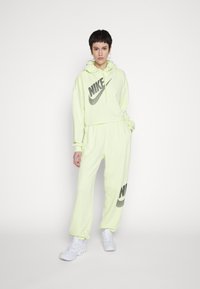 Felpa Nike gialla neon con logo nero, abbinata a pantaloni jogger coordinati con il logo, entrambi in tessuto morbido e testurizzato. Sneakers bianche completano il look.