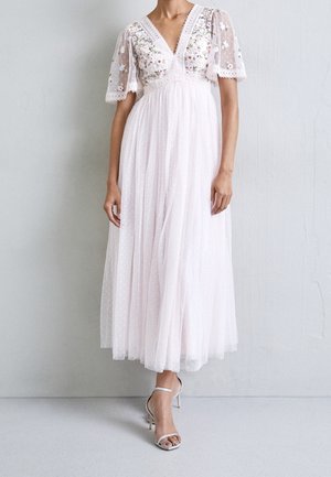 Mujer con un vestido blanco hasta el tobillo con mangas cortas transparentes bordadas con flores y falda plisada, combinado con tacones blancos con tiras en el tobillo.