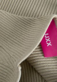 Beige ribbelstof met een zachte textuur; de gevouwen rand onthult een roze label met witte tekst. Aangegeven merknaam zichtbaar op het label.