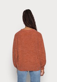 Pull en maille couleur rouille avec un col rond, des épaules tombantes et des manches longues légèrement bouffantes. Le tissu a une texture douce.