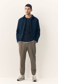 Navy jacket with a collar, layered over a dark shirt with a white trim, paired with beige trousers and gray sneakers. Smooth fabric textures.