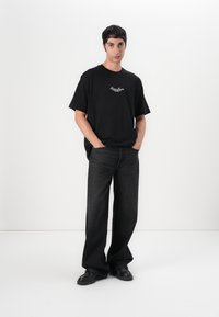 T-shirt en coton noir avec logo sur le devant, associé à un jean large gris foncé. Le mannequin porte des chaussures noires, se tenant devant un fond blanc.