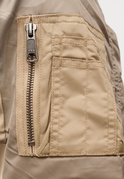 Τσέπη cargo από χακί ύφασμα με ασημί φερμουάρ YKK, διαθέτει ενισχυμένα ράμματα και υφή  επιφάνειας. Σχεδιασμένη για ανθεκτικότητα και λειτουργικότητα.