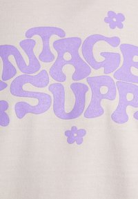 Krämfärgad tyg med djärv, vågig lila text som säger "VINTAGE SUPPLY", accentuerad med små lila blomformer.