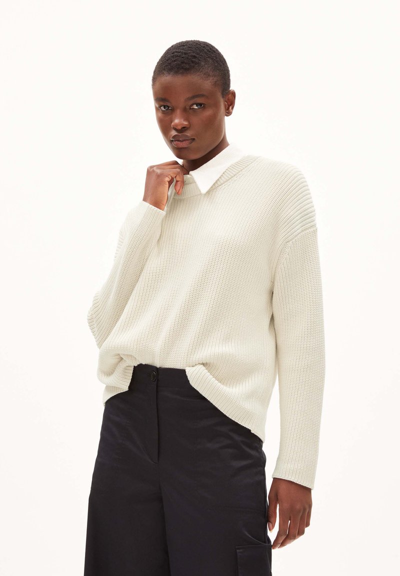 Hellcreme Strickjacke mit einem gerippten Muster und einem weiten, lockeren Schnitt, kombiniert mit dunkelblauen Hosen. Besitzt einen klassischen weißen Kragen.