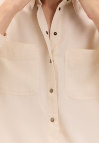 Chemise beige à boutons en tissu doux, dotée de deux poches poitrine et de boutons en métal. Design simple avec une coupe décontractée.