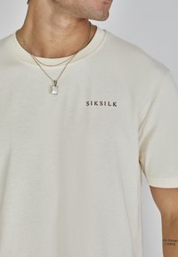 T-shirt à manches courtes de couleur crème avec une texture lisse et un logo subtil "SIKSILK" en brun foncé sur le devant. Chaînes superposées avec un pendentif.