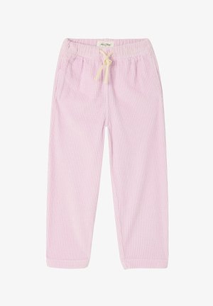 Mladé dievča s dlhými tmavými vlasmi, ktoré má na sebe béžové tričko s farebným logom, svetloružové nohavice a hnedé nazúvacie topánky, stojí v interiéri.