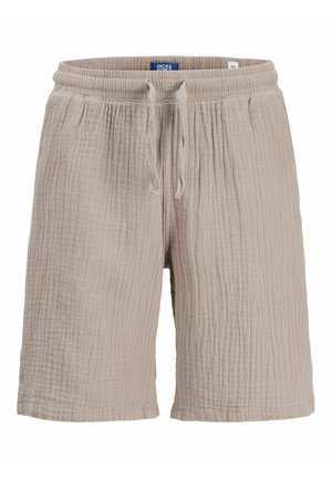 Chłopiec z kręconymi włosami, ubrany w koszulę na krótkim rękawie z abstrakcyjnym wzorem, beżowe szorty, białe skarpetki i beżowe buty, stojący na jednolitym tle.