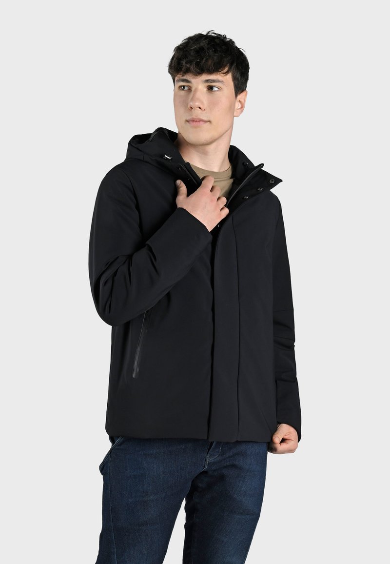 Giacca nera con cappuccio caratterizzata da una texture liscia, tasche con zip e un design elegante, indossata sopra una camicia chiara e jeans in denim.