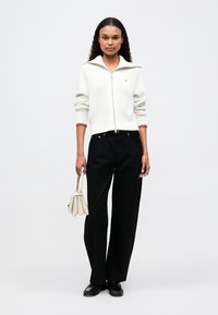 Biely rebrovaný sveter na zips s červenými a modrými akcentmi, skombinovaný s čiernymi širokými nohavicami a malou krémovou kabelkou.